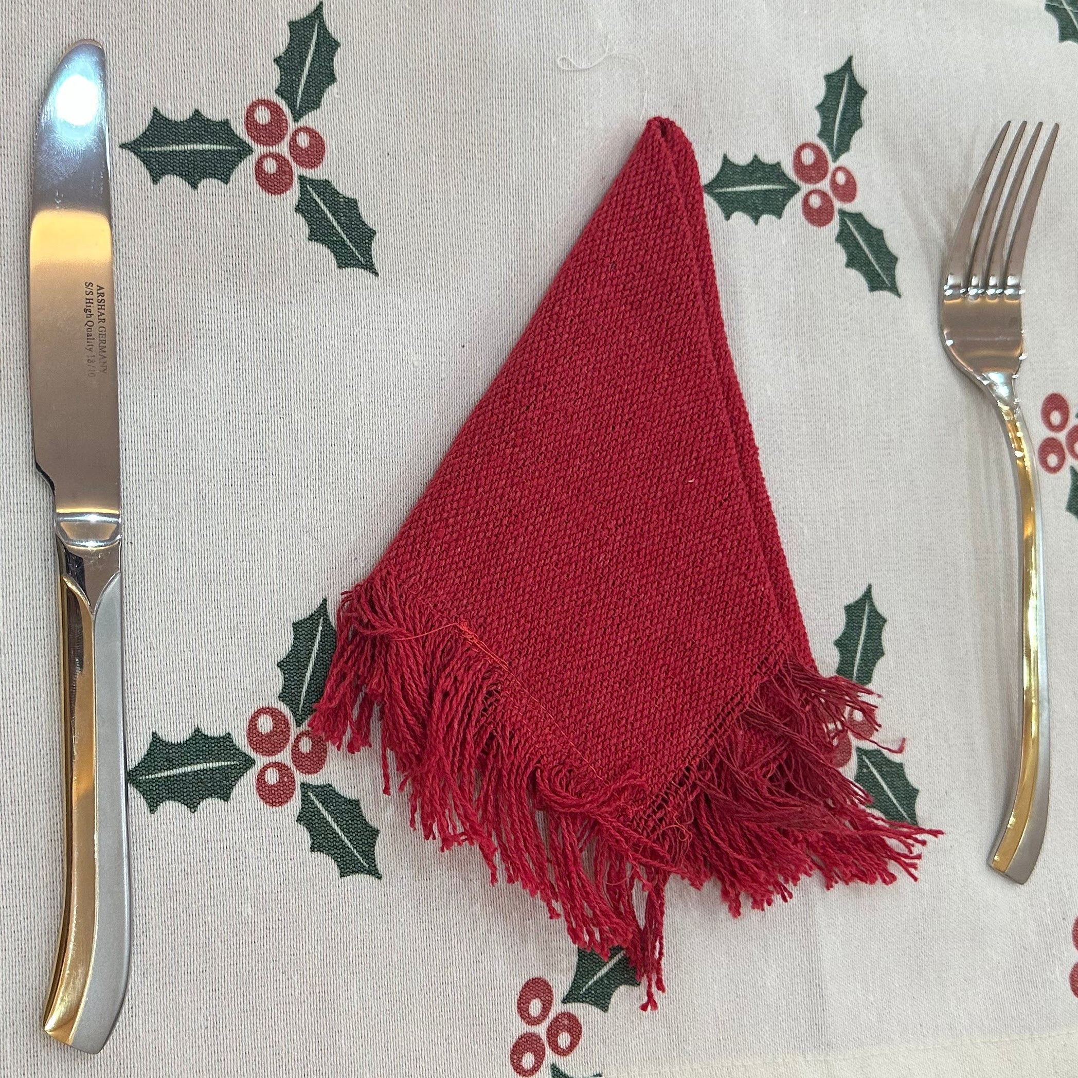 Decorative place mat with holly design, red napkin, and silverware on a red tablecloth.